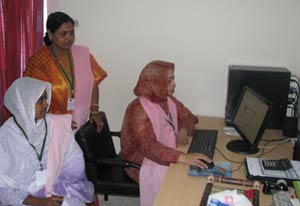 Three Bangladeshi women gather around a desktop computer, one seated using the mouse and keyboard, others behind her