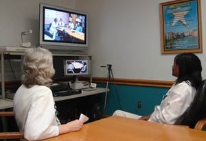 Two women seated at conference table, looking at teleconferencing equipment, with a screen that shows another conference room