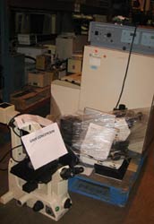 Large piles of scientific equipment including large microscopes and large metal box shapes, wrapped partially in clear plastic