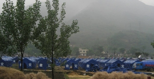 a photo of a refugee camp. Hundreds of blue tents are set up in a field with misty mountains behind them. Photo by Ma Hong.