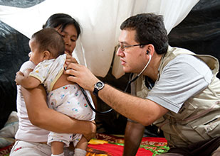 Doctor puts stethoscope on baby's back while mother holds the baby, all seated on ground, tarps in background