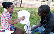 A female researcher in Uganda, seated across from a male research subject, points to low-literacy materials