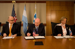 PHOTO: Seated at a long table Argentine Ambassador Héctor Timerman watches as NCI Director Dr. John E. Niederhuber and Urugayan Minister of Public Health Dr. Mária Julia Muñoz, sign papers.